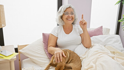 Poster - A smiling mature woman in glasses gesturing an idea while petting a dog in a cozy bedroom setting.