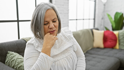 Wall Mural - A pensive, grey-haired woman sits thoughtfully in a cozy, modern living room, reflecting on mature adulthood.