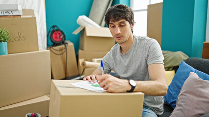 Sticker - Young hispanic man writing on document sitting on sofa at new home