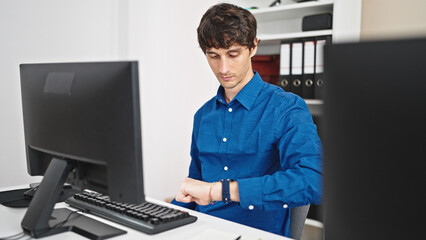 Wall Mural - Young hispanic man business worker using computer looking watch with serious face at the office