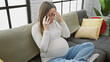 A pregnant hispanic woman sits on a couch at home, looking stressed while talking on the phone.