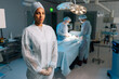 © dikushin - Portrait of serious African-American female nurse in surgical uniform standing posing looking at camera in operating theater. Multiethnic team of surgeons performing operation on blurred background.