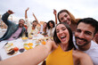© CarlosBarquero - Excited young pretty woman taking group selfie with joyful friends on rooftop. Cheerful people poses making smiling faces to camera outdoor gathered sitting table at lunch raising happy arms for photo