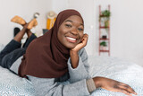 Fototapeta  - Portrait of young trendy african woman in muslim headscarf smiling at camera while lying on bed