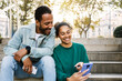 © Xavier Lorenzo - Two young latin american people using smart phone together sitting outdoors. Cheerful african american friends laughing while looking at cellphone screen. Social media concept. Copy space.