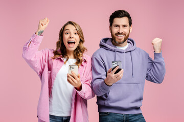 Portrait of overjoy hipster couple, man and woman wearing stylish colorful clothes, holding mobile phone, gesturing, win money, looking at camera isolated on pink background. Online technology concept
