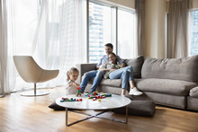 Carefree Couple Relaxing On Sofa, Little Daughter Play Wooden Cubes Seated At Table On Floor, Family Spend Pastime In Modern Apartment In Skyscraper Building. Leisure, New Home, Bank Loan, Tenancy