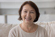 © fizkes - Positive mature older retired woman home head shot portrait. Female pensioner sitting on sofa, looking at camera with happy smile, enjoying retirement, talking on video conference call