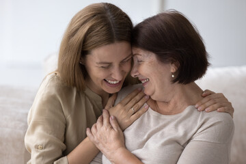Wall Mural - Joyful senior mom and adult daughter hugging, chatting, laughing at home, celebrating mothers day. Young woman embracing elder mum with closed eyes, face touches