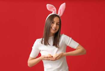 Wall Mural - Pretty young woman with bunny ears and Easter eggs on red background