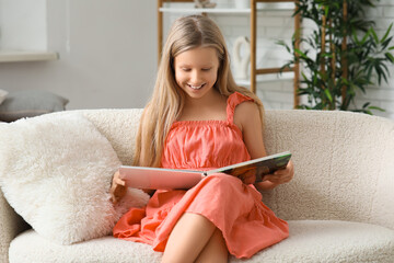 Poster - Cute little girl reading book while sitting on sofa at home