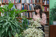 A girl with a phone in her hands stands between shelves with books and a shelf with flowers. The library is decorated in a modern style for a cozy relaxation area