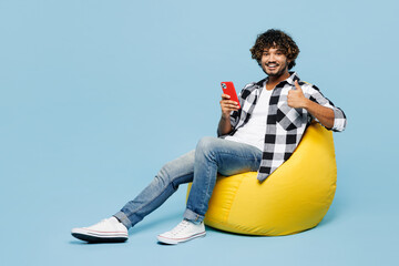 Wall Mural - Full body young Indian man wears shirt white t-shirt casual clothes sit in bag chair hold in hand use mobile cell phone show thumb up isolated on plain light blue cyan background. Lifestyle concept