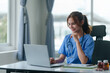 © amnaj - Friendly female nurse in blue scrubs seated at a desk, working on a laptop with medical charts and a stethoscope..