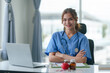 © amnaj - Female nurse in scrubs smiling confidently while seated at a desk with a laptop and medical equipment.