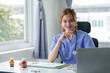 © amnaj - Friendly female nurse in blue scrubs seated at a desk, working on a laptop with medical charts and a stethoscope..