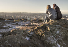 Two Women Sitting Relaxing On Top Of Rock Over The Mountain And Enjoying Looking At Scenic View Of Cityscape. Two Female Sitting Down Enjoying The Amazing View From The Mountain Top Of Salisbury Crags