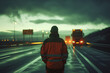 Man in reflective jacket facing away towards trucks on a highway at dawn