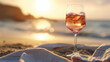 A glass of rosé wine on a cloth at the sandy beach during sunset, with the sun disappearing on the horizon, casting its reflection on the sea.