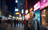 Fototapeta Konie - Joyful young muslim woman in hijab listening to music through headphones connected to cell phone while walking around the city center.