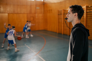 Wall Mural - Side view of a trainer on court using whistle while boys practicing basketball.