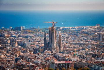 Panoramic view of Barcelona featuring the unfinished Sagrada Familia with its spires and cranes, amidst a grid of streets and diverse architecture, with the Mediterranean Sea in the backdrop.