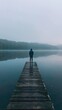Person standing at end of dock, misty lake, serene atmosphere, wooden pier, reflective water, foggy environment, solitude, contemplative scene, blue tones, vertical orientation
