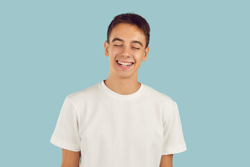 Portrait of a young smiling cheerful boy student wearing casual white t-shirt standing with closed eyes isolated on a studio blue background. People emotions and lifestyle concept.