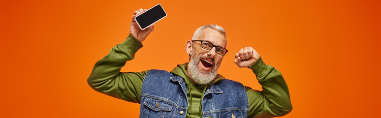 Wall Mural - jolly handsome mature man in green hoodie and denim vest holding phone and looking at camera, banner