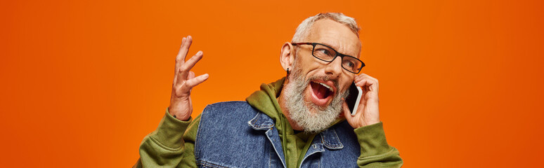 Wall Mural - handsome jolly mature man in green hoodie and denim vest with glasses talking by phone, banner