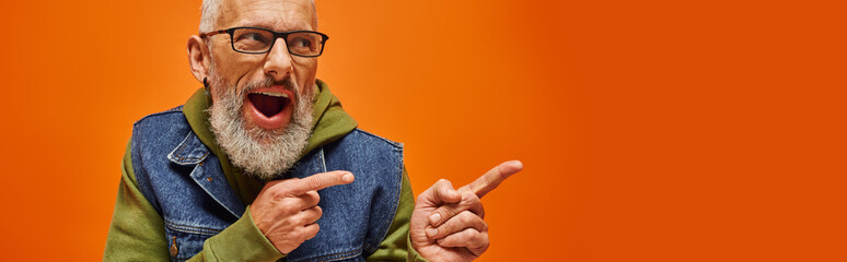 Wall Mural - joyful handsome mature man in trendy denim vest posing on orange backdrop and pointing away, banner