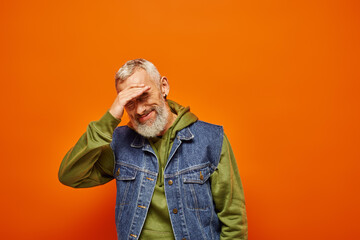 Wall Mural - joyful good looking mature man in vivid attire with gray beard smiling at camera on orange backdrop