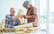 © Ann Rodchua - Two senior mixed race diverse happy retired old male woodworkers or carpenters wearing check shirts, creating DIY wooden car toy together for decoration and furniture. Hobby, Retirement Concept.