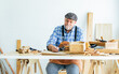 © Ann Rodchua - Senior Caucasian happy retired old male woodworker or carpenter smiling, wearing check shirt with apron, vintage hat, creating DIY wooden mini figure toy for decoration as hobby after retirement.