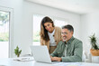 © insta_photos - Smiling middle aged senior man working on computer sitting at table with wife standing nearby in living room. Happy mature older couple using laptop technology at home. Authentic candid shot.