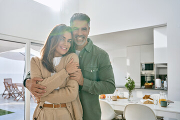 Wall Mural - Happy romantic mature older Latin man and woman in their 50s, smiling affectionate loving middle aged couple hugging looking at camera standing together in modern kitchen at home. Portrait.