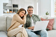 © insta_photos - Happy middle aged couple using laptop relaxing on couch at home. Smiling mature man and woman looking at computer watching video, browsing or shopping in ecommerce store sitting on sofa in living room
