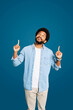 © Vadim Pastuh - A handsome young Brazilian man with a pleasant smile points upwards with both hands, looking upwards with an expression of optimism, against a calm blue background. Concept of advertising