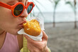 Young blond woman at seaside eating hot palatable arancini (deep fried rice balls with meat). Typical Sicilian street food. Close up.