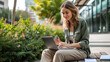 © Studio Nova - young woman is focused on working on her laptop while sitting at a wooden table outdoors, with plants around and modern buildings in the background