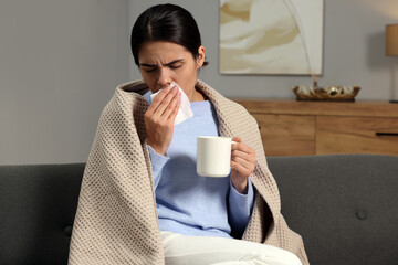 Poster - Woman with tissue and cup of drink coughing on sofa at home. Cold symptoms