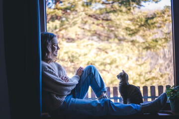 Wall Mural - Young smiling woman in a warm sweater with a cute gray cat on the windowsill at home