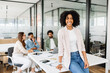 © Vadim Pastuh - Charming smart Brazilian woman, a female office employee, stands with easy confidence, her team is in the background, embodying a relaxed yet productive meeting atmosphere