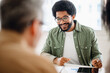 © Vadim Pastuh - African-American man with a warm smile, wearing glasses and a green casual shirt, is engaged in conversation with an out-of-focus colleague, suggesting an approachable and friendly office discussion