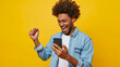 an exuberant young person with curly hair, holding a smartphone and seemingly shouting or cheering with a bright yellow background