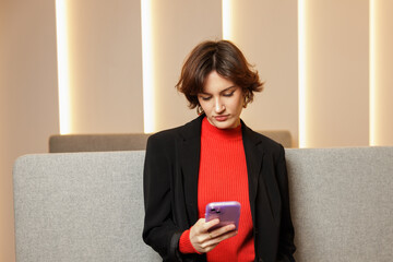 Young gen z woman holding a mobile phone. Female in red turtleneck and black formal blazer. Businesswoman in office environment. Colorful and joyful attitude