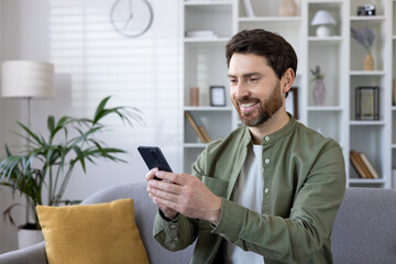 Happy bearded man enjoying using cellular phone while sitting on couch in living room. Careless caucasian guy looking at digital gadget screen while scrolling news feed on local websites indoors.
