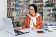 © Liubomir - Inspired female student sitting in library by desk with laptop and books while doing notes by hand on paper sheet. Interested lady watching online webinar lecture and making remarks for memorization.