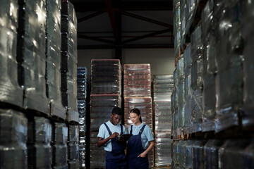 Poster - Two young factory workers in coveralls standing among stacks of packages with bottled water and discussing data on tablet screen