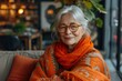 A joyful senior woman sits indoors, adorned in vibrant orange shawl and glasses, radiating warmth and contentment with her genuine smile and bold fashion choice